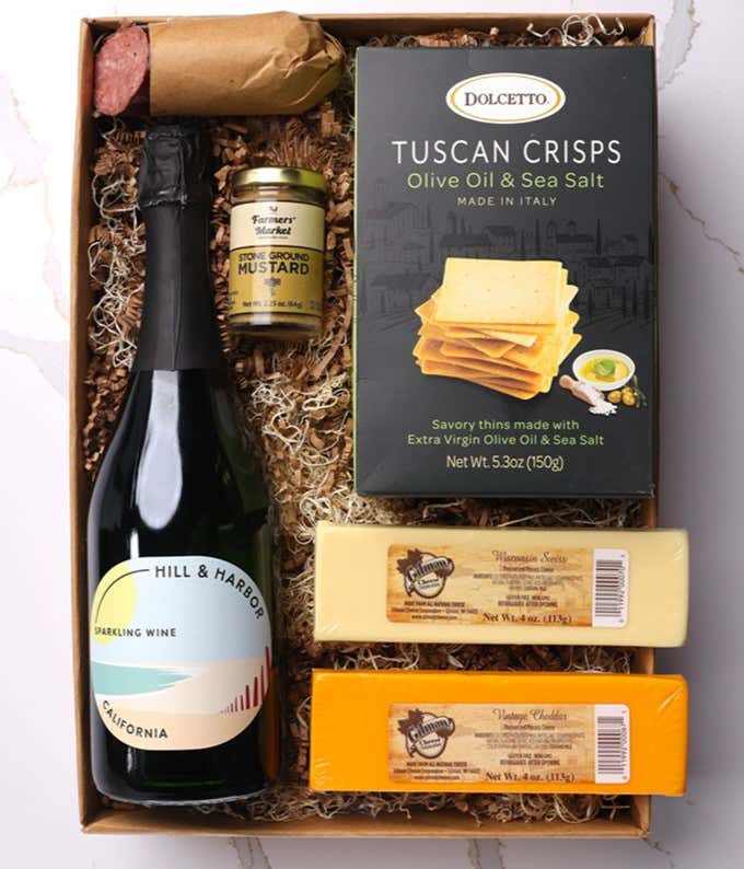 A gift box with Tuscan crisps, salami, cheeses, mustard, and a bottle of Hill & Harbor sparkling wine. Surrounding the box are plates with cheese, salami, mustard, and two glasses of sparkling wine.