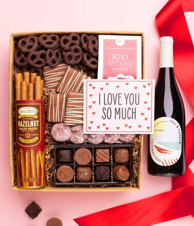 A gift box with a bottle of red wine, chocolate pretzels, chocolate graham crackers, red velvet cookies, hazelnut wafer rolls, an 8 piece box of chocolates and an I Love You card.