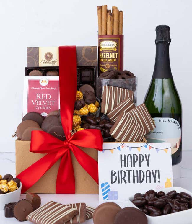 A gift box overflowing with treats including an assortment of chocolate-covered items like oreoes, almonds graham crackers and pretzels along with red velvet cookies, hazelnut wafers, an 8 piece box of chocolates, a bottle of sparkling wine and a Birthday