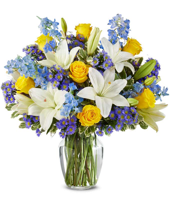An elegant floral arrangement featuring white Asiatic lilies, yellow roses, blue delphinium, and purple Monte Casino, optionally arranged in a glass vase. This vibrant mix offers a stunning and colorful display.