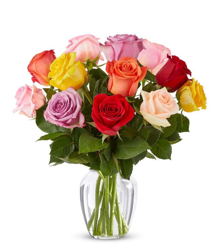 A bouquet of multicolored roses in a clear vase, featuring red, pink, orange, yellow, peach, and lavender blooms, arranged with green leaves for a vibrant and cheerful display.