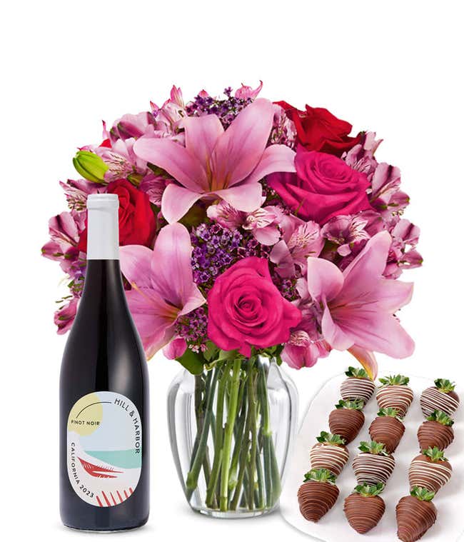 A mixed bouquet of pink and purple flowers in a clear glass vase is displayed with a bottle Hill &amp; Harbor Pinot Noir and 12 chocolate-covered strawberries.