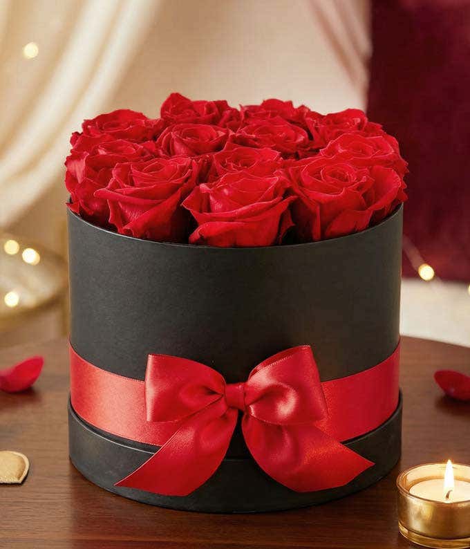 Arrangement of vibrant red roses in a round black box, adorned with a red satin ribbon tied into a bow.