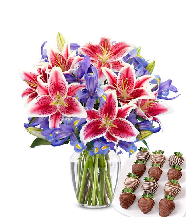 Clear glass vase filled with a vibrant bouquet of hot pink stargazer lilies and deep violet-blue irises, paired with a plate of swizzled chocolate-covered strawberries drizzled in dark and white chocolate.