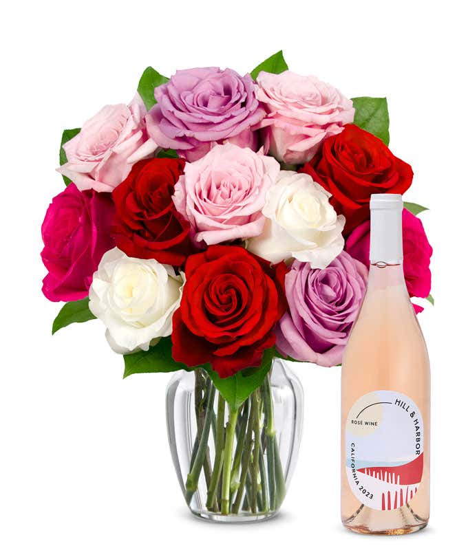 One dozen pink, purple and red roses arranged in a clear glass vase sits beside a bottle of Hill & Harbor California Rosé.