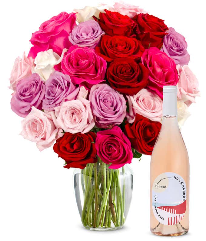 Two dozen pink, purple and red roses arranged in a clear glass vase sits beside a bottle of Hill & Harbor California Rosé.