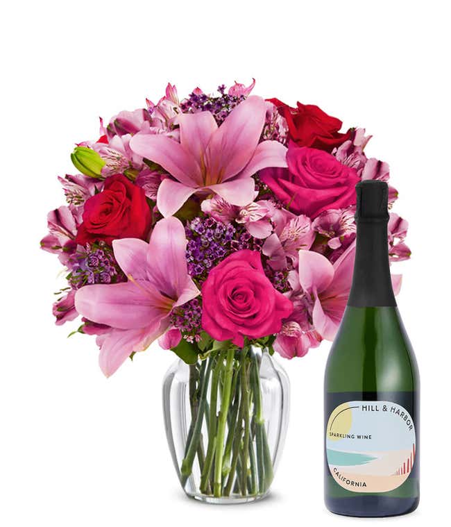 A bouquet of pink lilies, pink roses, and greenery arranged in a clear glass vase sitting next to a bottle of Hill &amp; Harbor California Sparkling Wine.