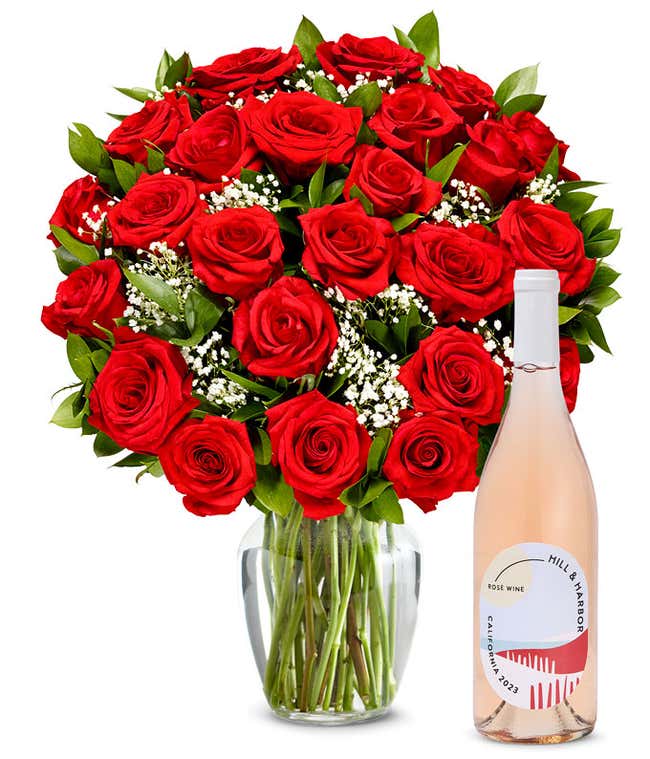 A lush bouquet of red roses, with deep velvet petals, nestled in a clear vase among tiny white blossoms and green leaves, complements a sleek rose wine bottle, vintage 2023 from California, radiating elegance and romance.
