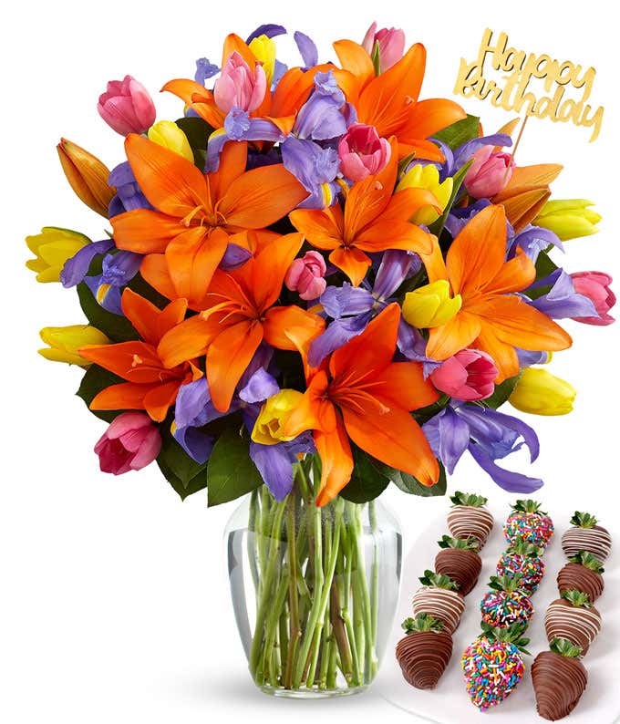 Deluxe bouquet of orange lilies, purple irises, and multicolored tulips in a clear glass vase with a gold Happy Birthday pick, paired with a plate of swizzled and rainbow sprinkle chocolate-covered strawberries.