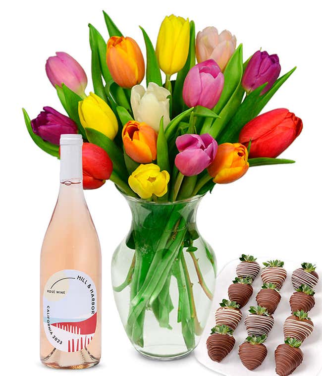 A clear glass vase filled with a 15-stem assortment of multi-colored tulips paired with a bottle of ros&eacute; wine and 12 chocolate-covered strawberries.