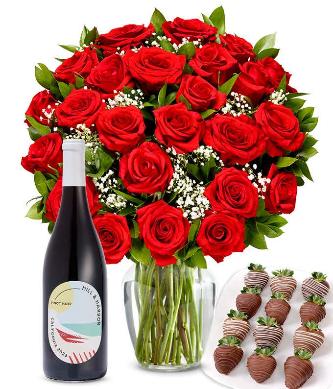 A bouquet of 2 dozen red roses in a clear glass vase is displayed beside a bottle of Hill & Harbor Pinot Noir and an arrangement of 12 chocolate-covered strawberries.