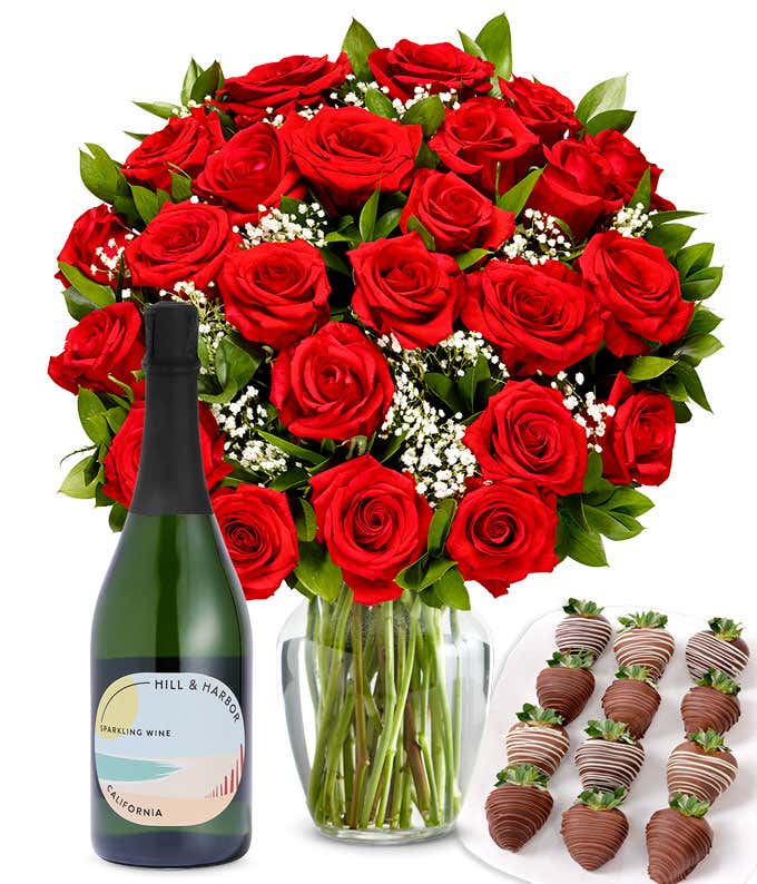 A bouquet of 2 dozen red roses in a clear glass vase is displayed beside a bottle of Hill & Harbor Sparkling Wine and an arrangement of 12 chocolate-covered strawberries.