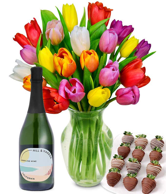 A clear glass vase filled with a 20-stem assortment of multi-colored tulips paired with a bottle of sparkling wine and 12 chocolate-covered strawberries.