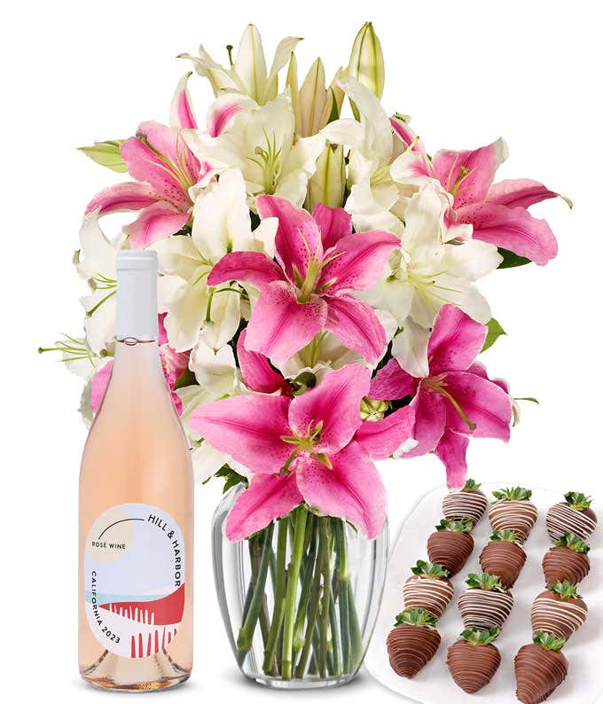 A bouquet of pink and white lilies arranged in a clear glass vase is paired with a bottle of Hill & Harbor Rosé and 12 chocolate-covered strawberries.