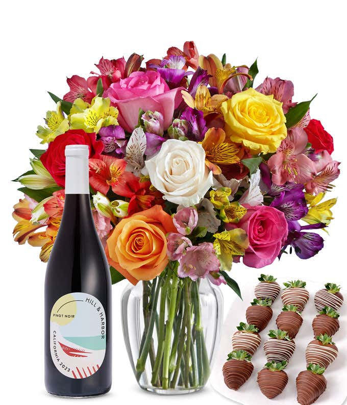 A bouquet of colorful roses and alstroemeria in a clear vase is displayed beside a bottle of Hill & Harbor Pinot Noir and 12 chocolate-covered strawberries.
