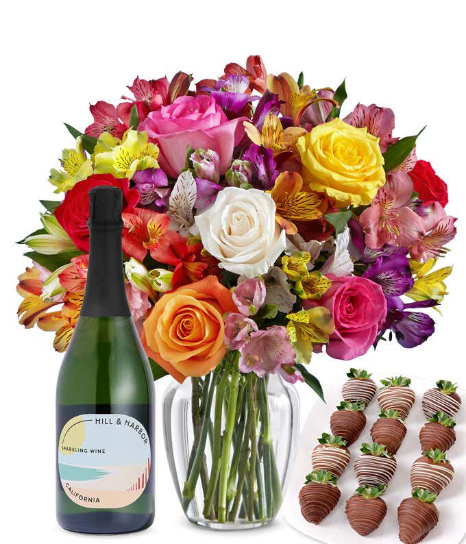 A bouquet of colorful roses and alstroemeria in a clear vase is displayed beside a bottle of Hill & Harbor Sparkling Wine and 12 chocolate-covered strawberries.