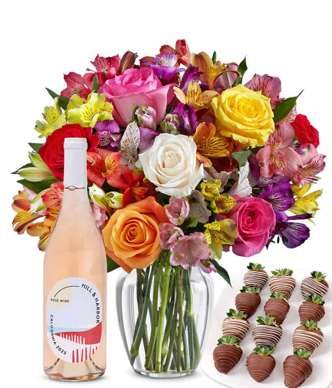 A bouquet of colorful roses and alstroemeria in a clear vase is displayed beside a bottle of Hill &amp; Harbor Ros&eacute; and 12 chocolate-covered strawberries.