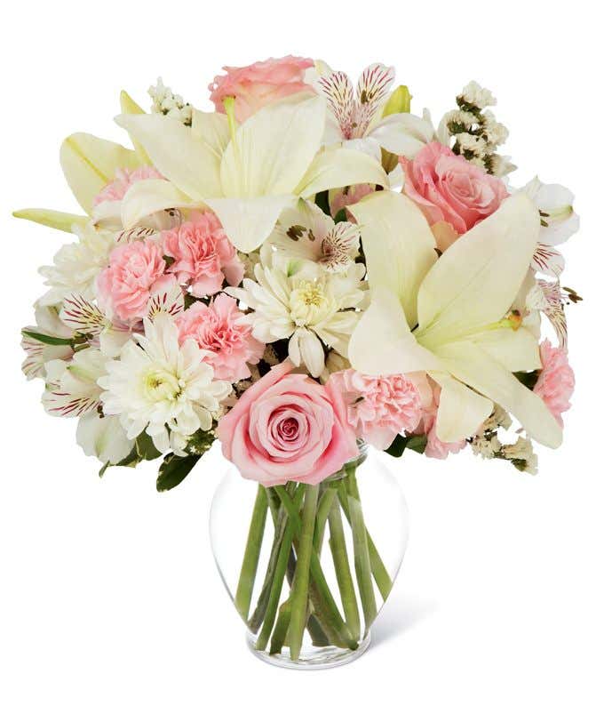 Short, round arrangement of white lilies, mums, and alstroemeria, and pink roses and carnations, in a glass vase