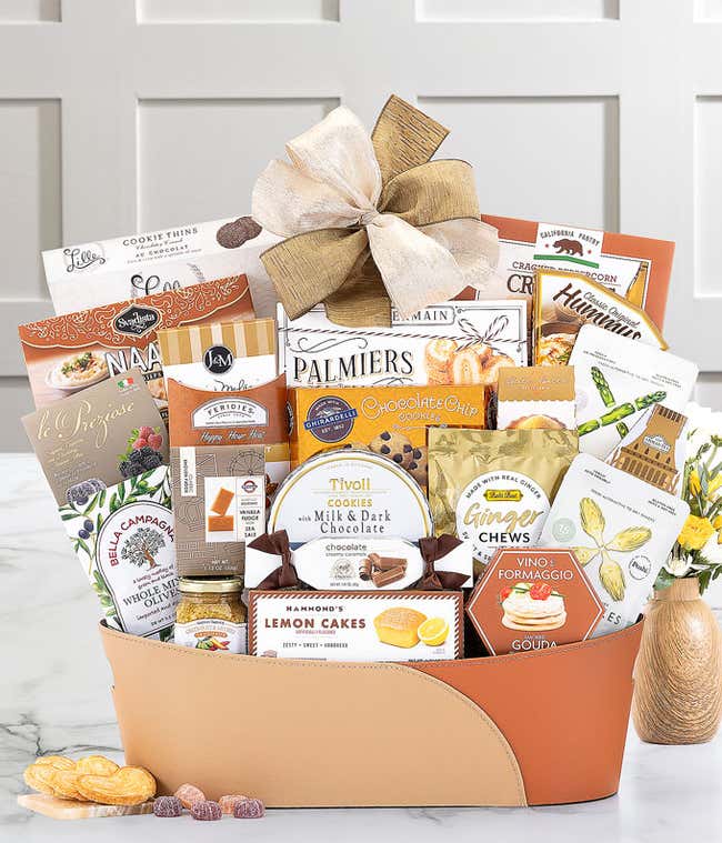 Tan and brown gift basket with a gold bow, filled with assorted snacks including cookies, chocolates, tea, hummus, olives, naan crisps, and cheese spread, displayed on a marble surface with a small flower vase.