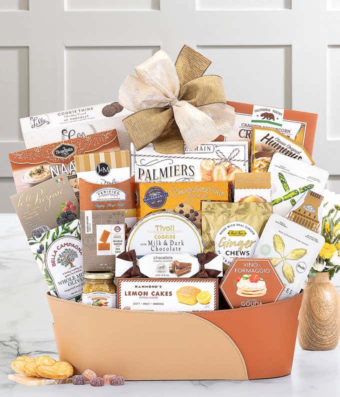 Tan and brown gift basket with a gold bow, filled with assorted snacks including cookies, chocolates, tea, hummus, olives, naan crisps, and cheese spread, displayed on a marble surface with a small flower vase.