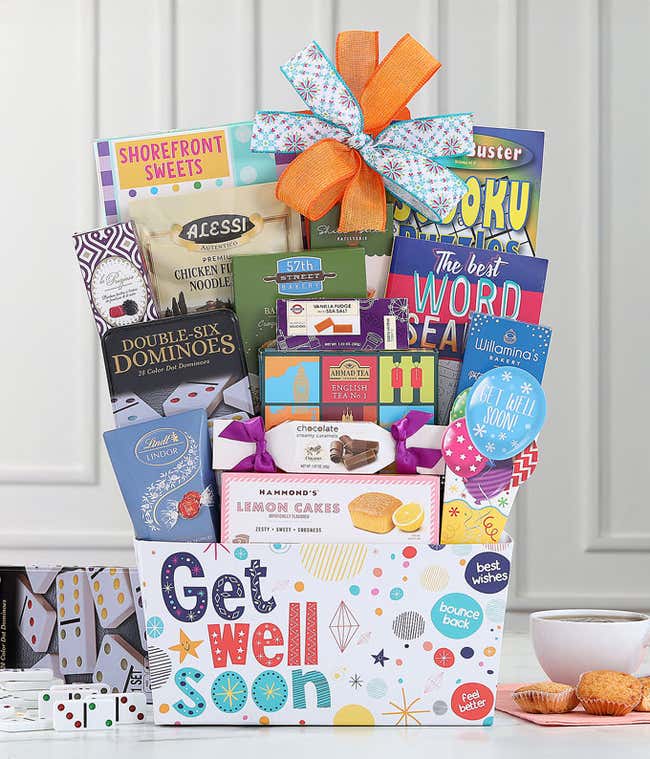 Get well soon gift basket with an orange and blue bow, filled with Ahmad English Tea, baked crackers, soup mix, sweet treats, puzzle books, and a dominoes set, displayed with tea and snacks.