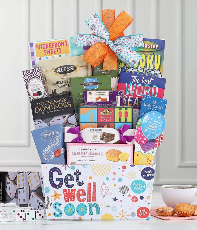 Get well soon gift basket with an orange and blue bow, filled with Ahmad English Tea, baked crackers, soup mix, sweet treats, puzzle books, and a dominoes set, displayed with tea and snacks.