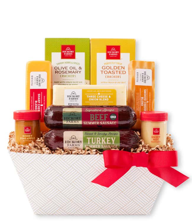 Gift basket with summer sausages, cheeses, crackers, and mustards in a white patterned container with a red bow. A serving board in front displays sliced sausage, cheese, and mustard for dipping.