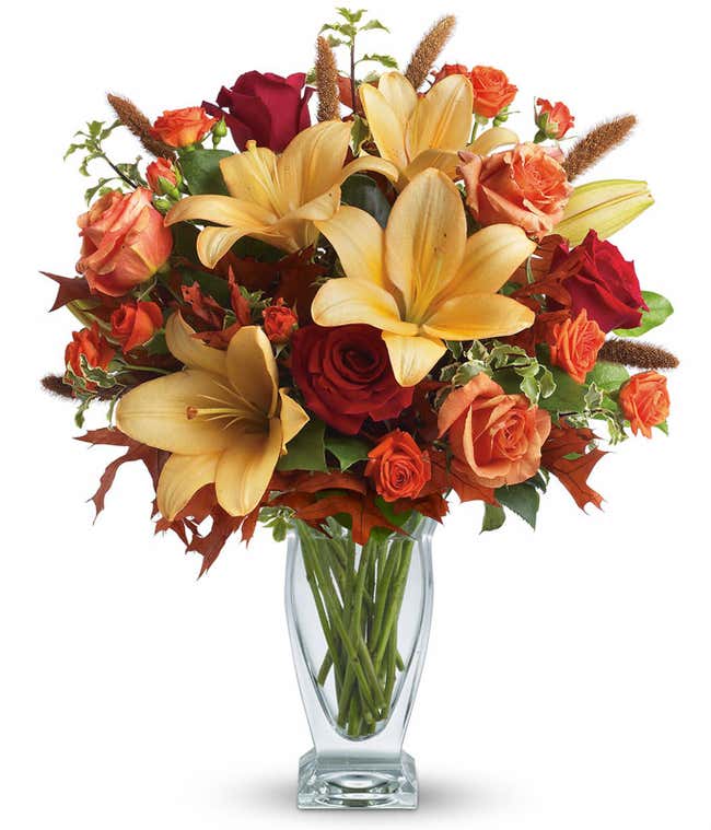 A stunning floral arrangement featuring orange roses, red roses, orange Asiatic lilies, and dried leaves, all elegantly presented in a glass vase.