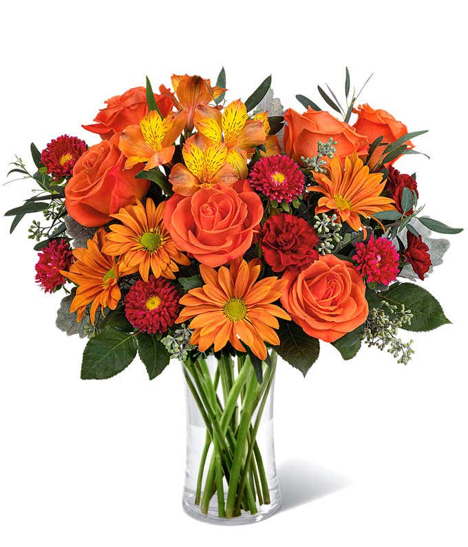 Vibrant fall bouquet with orange roses, orange daisies, red carnations, pink Matsumoto asters, yellow-orange alstroemeria, and assorted greenery in a clear glass vase.