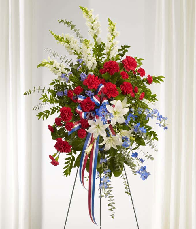 White lilies, white snapdragons, blue delphinium, red carnations with multicolored patriotic ribbon.