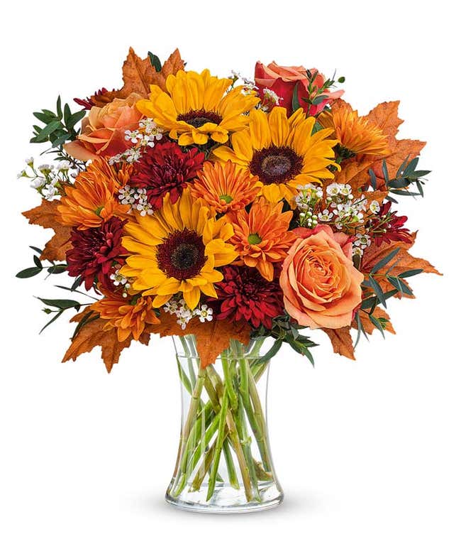 A cheerful floral arrangement featuring orange roses, red cushion spray mums, bronze daisy spray mums, and bright yellow sunflowers, accented with seasonal floral greenery, and presented in a clear glass vase..