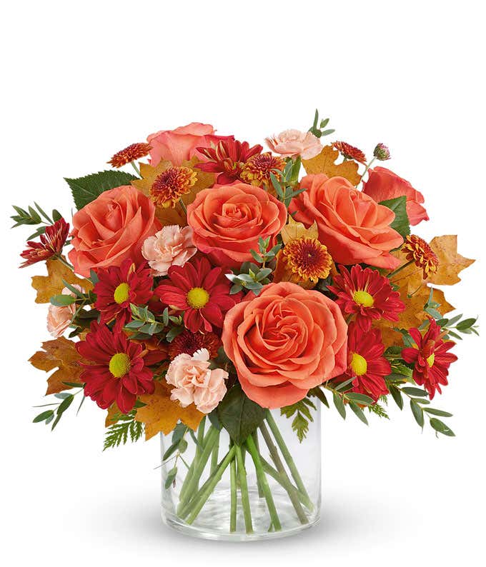 A vibrant arrangement with orange roses, miniature peach carnations, bronze button spray mums, red daisy spray mums, and seasonal greenery in a clear glass vase.