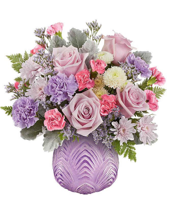 Soft pastel floral arrangement featuring lavender roses, lavender carnations, pink miniature carnations, lavender cushion spray chrysanthemums, white button spray chrysanthemums, dusty miller, and lush greenery in a textured lavender glass vase.