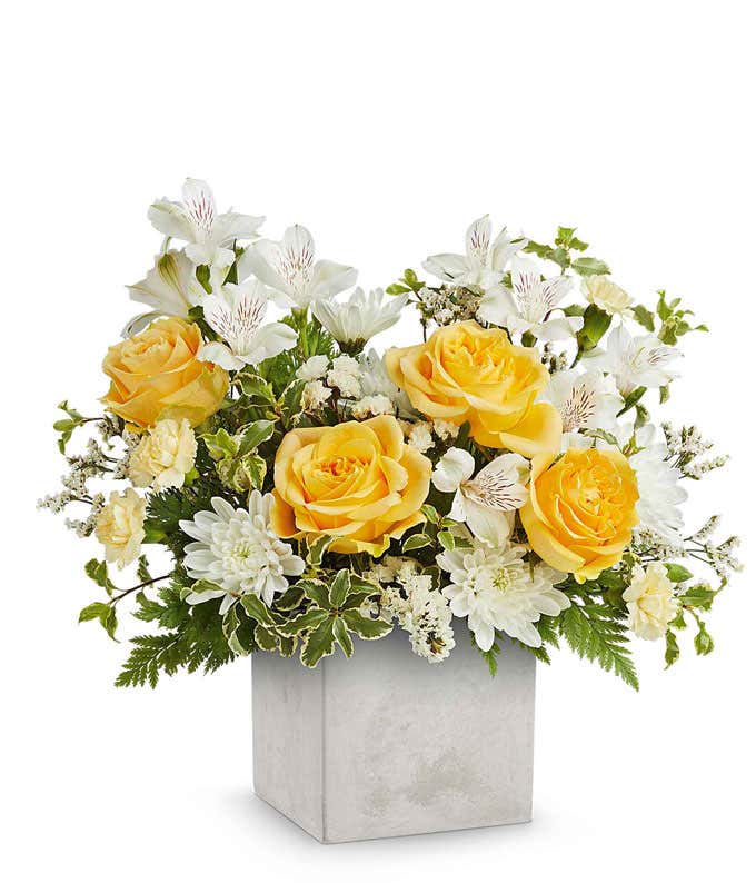 A floral arrangement in a square gray container featuring yellow roses, white alstroemeria, white carnations, white poms, ivy, and seasonal greenery.