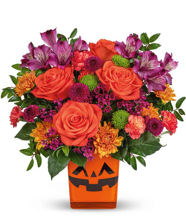 Halloween-themed flower arrangement featuring orange roses, purple alstroemeria, green button spray chrysanthemums, and assorted mums in a bright orange jack-o'-lantern container.
