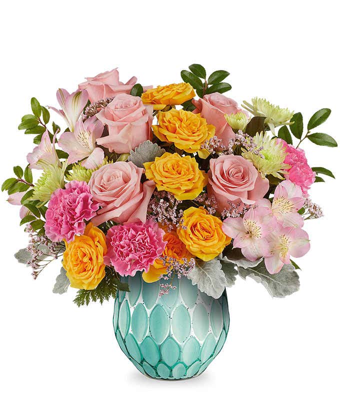 Pink roses, yellow spray roses, hot pink carnations, pink alstroemeria, green button spray chrysanthemums, pink waxflower, and assorted greenery arranged in a textured turquoise glass vase.