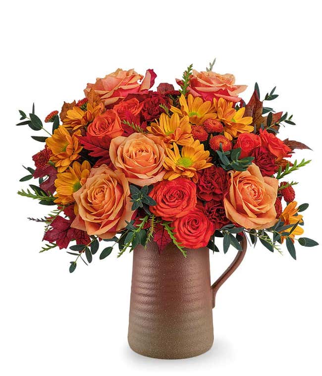 Fall flower arrangement with orange roses, orange spray roses, maroon carnations, brown button spray chrysanthemums, and autumn leaves in a brown ceramic pitcher.