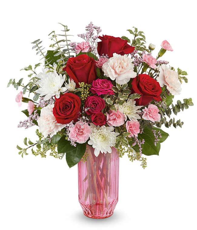 Valentine&rsquo;s Day floral arrangement featuring red roses, blush and pink blooms, white flowers, and lush greenery arranged in a pink glass vase.
