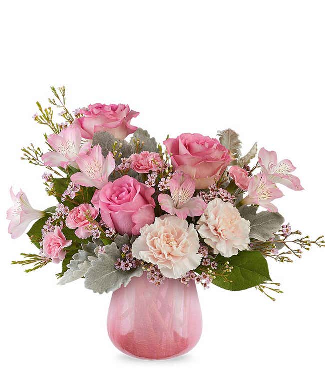 Romantic floral arrangement featuring soft pink roses, blush carnations, pale pink alstroemeria, silvery dusty miller, and lush greenery arranged in a pink glass vase.
