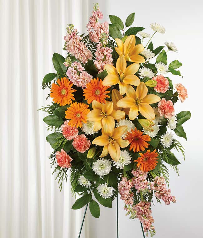 A standing funeral spray featuring Peach Asiatic Lilies, Orange Gerberas, Orange Carnations, Peach Stock, White Cushion Chrysanthemums, and Floral Greenery, offering a serene tribute of comfort and solace during a time of loss.