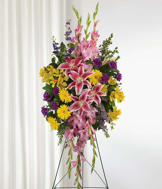 A standing funeral spray adorned with Pink Stargazer Lilies, Yellow Gerberas, Yellow Alstroemeria, Pink Gladioli, Purple Carnations, and Purple Larkspur, evoking a sense of grace, warmth, and reverence in remembrance of a beloved soul.