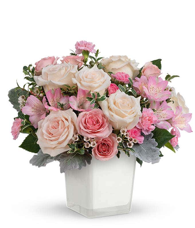A floral arrangement featuring crème roses, pink spray roses, pink alstroemeria, pink miniature carnations, white waxflower, and greenery in a white vase.