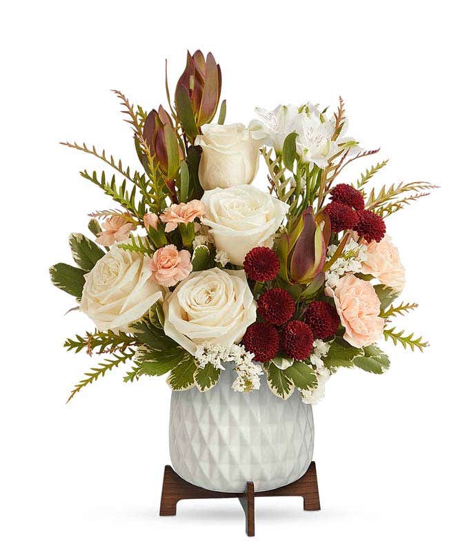 A bouquet of crème roses, white alstroemeria, red leucadendron, peach carnations, and burgundy button spray chrysanthemums in a white vase with a wooden stand.