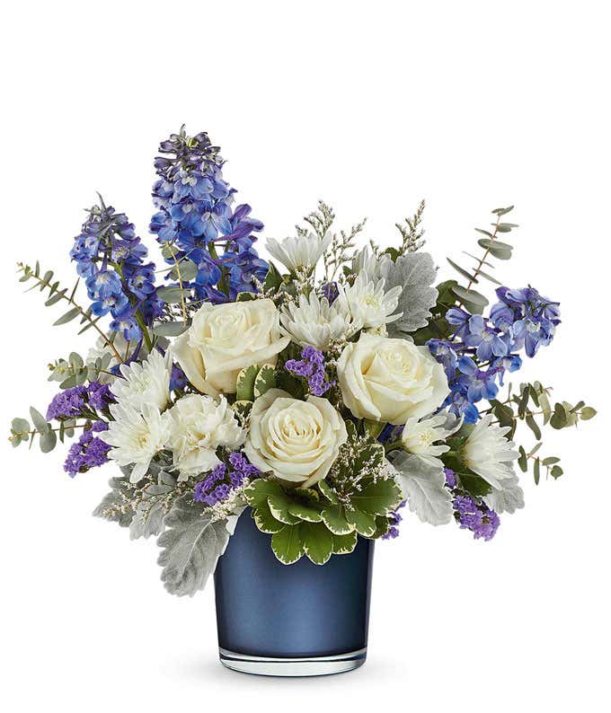 Elegant floral arrangement featuring white roses, white poms, deep blue delphinium, purple statice, dusty miller, silver dollar eucalyptus, and assorted greenery in a sleek navy blue glass vase.