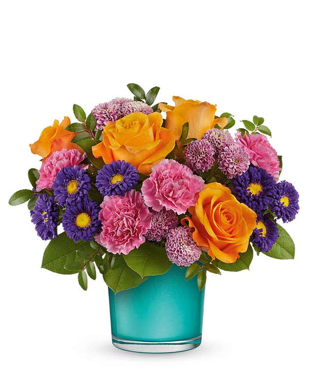 Vibrant floral arrangement featuring orange roses, pink carnations, purple asters, lavender poms, and lush greenery in a turquoise glass container.