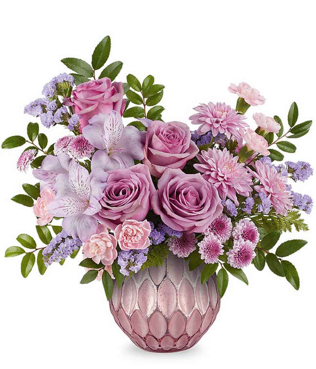 Lavender roses, lavender alstroemeria, miniature pink carnations, lavender button spray chrysanthemums, and lavender sinuata statice accented with greenery in a textured pink vase.