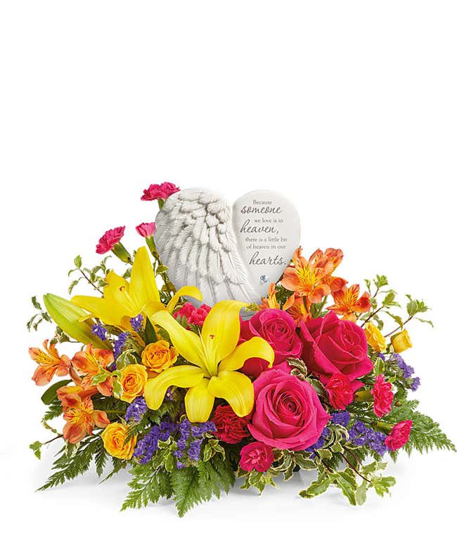 Sympathy flower arrangement with bright yellow lilies, hot pink roses, orange alstroemeria, miniature carnations, and assorted greenery, arranged around a ceramic heart-shaped keepsake with angel wings and an inscription about loved ones in heaven.