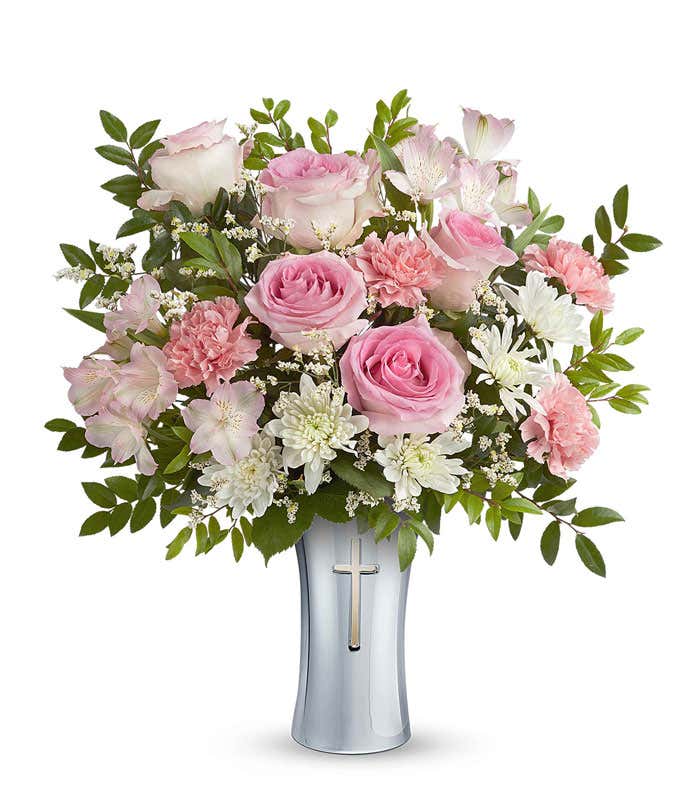 Floral arrangement with pink roses, pink alstroemeria, pink carnations, white cushion spray chrysanthemums, white limonium, and assorted greenery in a keepsake silver vase featuring a cross design.