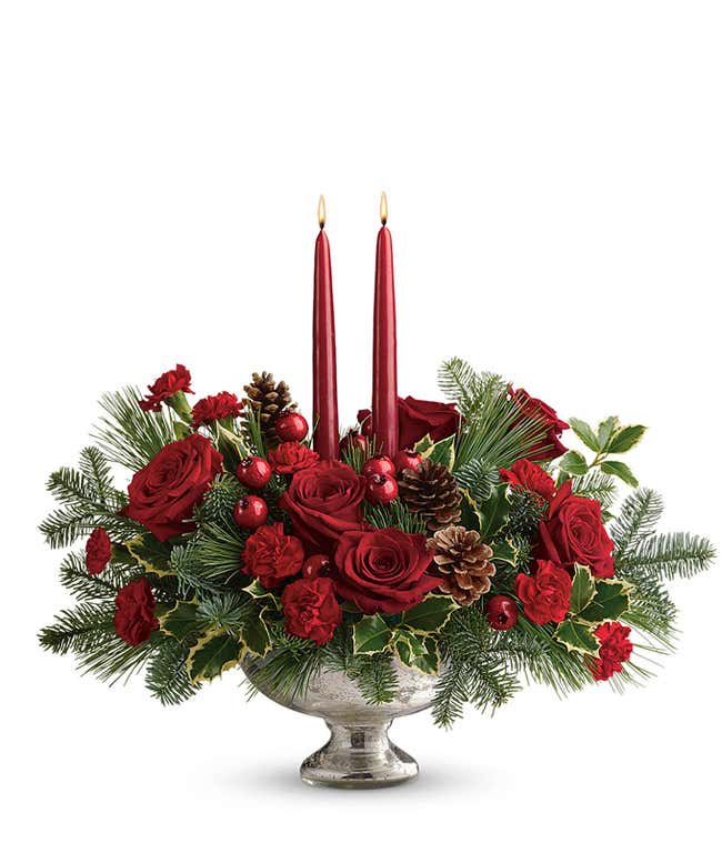 Holiday centerpiece featuring red roses, red miniature carnations, festive berry sprays, pinecones, evergreen greenery, and two red tapered candles arranged in a keepsake mercury glass bowl