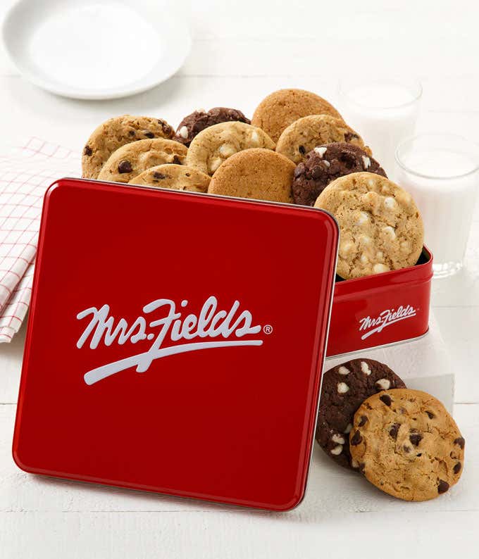  Red Mrs. Fields tin filled with chocolate chip cookies displayed with glasses of milk and striped straws on a white wooden surface.